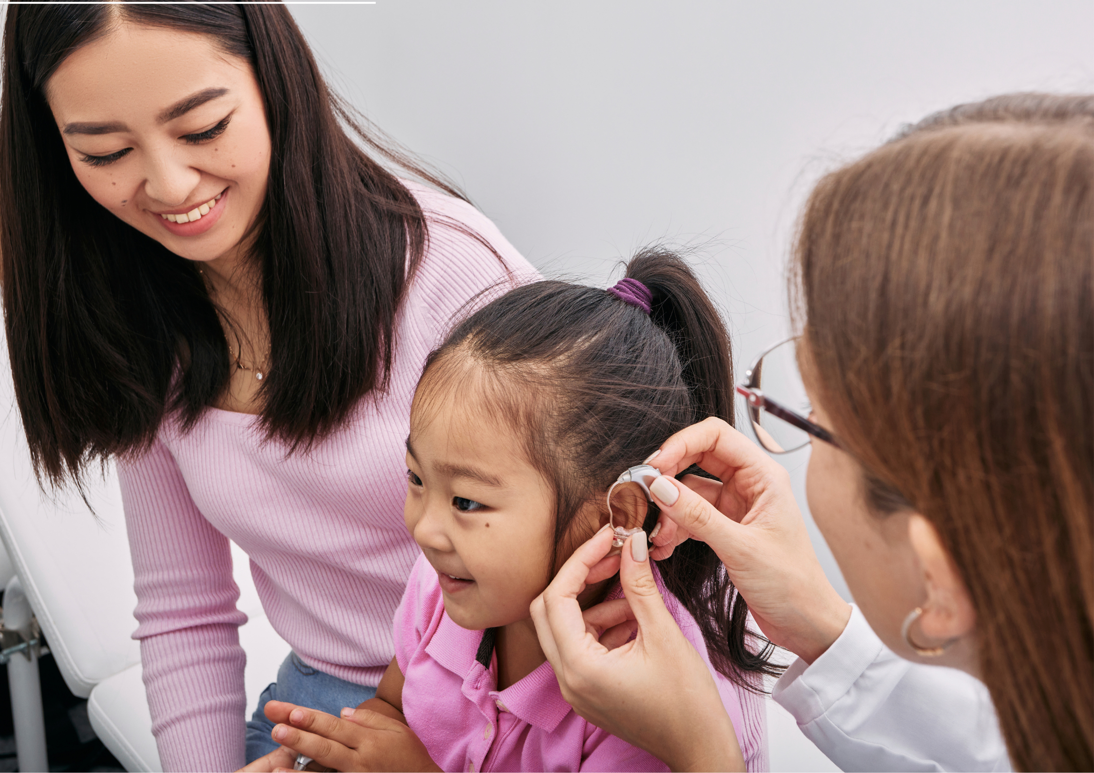 Pediatric hearing services: an audiologist examining a young girl's ear while her mother watches.