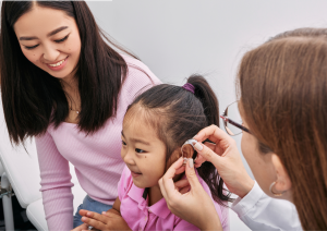 Pediatric audiologist fitting a small hearing aid device on a child as her mother looks on.